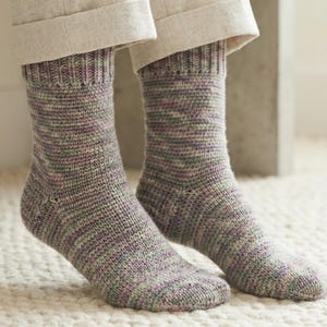 May include: A pair of knitted socks in shades of gray, green, and purple. The socks are worn with light-colored pants. The socks have a ribbed cuff and a striped pattern. The person is sitting on a concrete bench.
