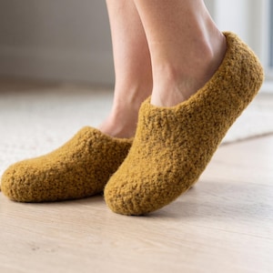 Könnte beinhalten: Ein Paar senfgelbe Hausschuhe mit einer strukturierten, filzartigen Optik. Die Hausschuhe verfügen über eine abgerundete Zehenpartie, einen tiefen Einstieg und ein Slipper-Design, das den Fuß bis zum Knöchel umschließt und einheitlich wirkt.