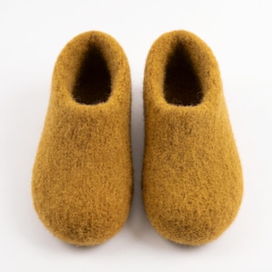 May include: A pair of mustard yellow felted wool slippers with a rounded toe and a slip-on design. The slippers feature a thick, textured surface and a deep opening, showcasing a solid, uniform color throughout the entire exterior construction.