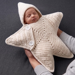 May include: A cream-colored, star-shaped crochet swaddle blanket with an integrated hood. The garment features a textured, open-stitch pattern and is secured at the front with two white circular buttons. The design forms a five-pointed star shape.