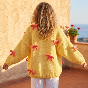 Könnte beinhalten: Ein gelber Strickpullover mit mehreren rosa Schleifen. Der Pullover hat lange Ärmel und eine lockere Passform. Die Schleifen sind gleichmäßig über Vorder- und Ärmel verteilt. Der Hintergrund ist eine hellbraune Wand und das Meer.