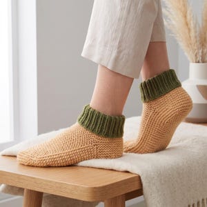 May include: A pair of ankle-length knit socks featuring a light tan main body with a textured stitch pattern and a contrasting dark olive green ribbed cuff. The socks are designed to sit just above the ankle bone with a snug, fitted construction.