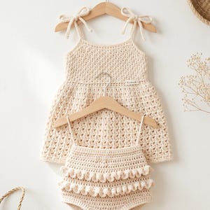 Pode incluir: Um conjunto de vestido e calcinha de bebê de crochê feito à mão, cor creme. O vestido apresenta alças amarradas e uma saia rodada. A calcinha tem um design com babados. Ambas as peças são exibidas em cabides de madeira.
