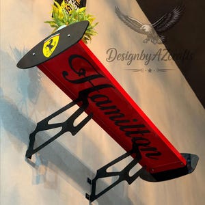 May include: A red automotive-style rear spoiler wall shelf featuring black support brackets. The top surface displays a yellow Ferrari logo shield. The word Hamilton is written in black cursive script across the red surface of the spoiler.
