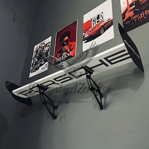 May include: A white Porsche-branded spoiler mounted on a gray wall, with black support brackets. The spoiler is positioned below several framed posters featuring automotive and music themes. The word "Porsche" is prominently displayed on the spoiler.