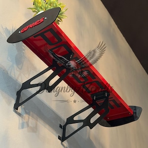 May include: A red and black car spoiler with the word "PORSCHE" in large black letters. The spoiler has a black top with the word "GRES" in red. The spoiler is mounted on a wall with black brackets.