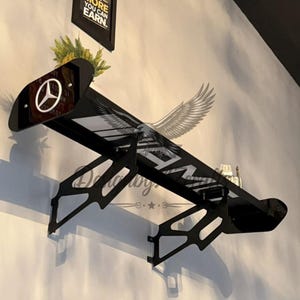 May include: Black wall-mounted shelf in the shape of a car spoiler. The shelf has a white and black design with the word "AMG" and a logo. The shelf is mounted on a light-colored wall.