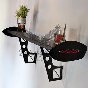 May include: Black wall shelf in the shape of a car spoiler, with a dark brown shelf surface. The shelf has a decorative eagle graphic and the text "GT3RS" in red. A small plant, perfume, and Eiffel Tower figurine are displayed on the shelf.