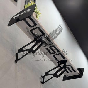 以下が含まれることがあります： ポルシェブランドのスポイラーデザインを特徴とする白黒の壁の装飾。デザインには、黒い翼に白い文字で「Porsche」という単語、そして「DesignbyArt」という単語とワシのグラフィックが含まれています。装飾は白い壁に取り付けられています。