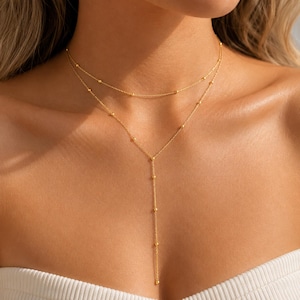 Könnte beinhalten: Eine goldfarbene, mehrlagige Halskette bestehend aus zwei zarten Ketten. Die obere Kette ist ein schlichtes Choker-Modell, während die untere Kette einen Y-förmigen Anhänger bildet. Beide Ketten sind mit kleinen, gleichmäßig verteilten Goldperlen besetzt.