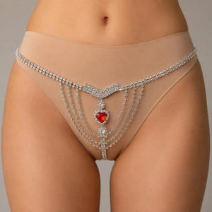 Peut inclure: Un string beige agrémenté d'une chaîne de corps décorative argentée ornée de strass transparents. Le design de la chaîne comprend un accent central en forme de V, plusieurs rangées drapées et un pendentif en strass rouge en forme de cœur.
