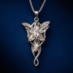 Può includere: Una collana con pendente color argento caratterizzata da un design floreale stilizzato con cristalli trasparenti taglio marquise disposti a raggiera. La montatura in metallo presenta intricate texture a foglia e un delicato nodo intrecciato.