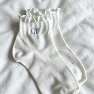 May include: A pair of white ankle socks with a ruffled trim at the top. The socks have a ribbed texture and a blue embroidered "CT" on the cuff. The socks are displayed on a white, textured surface.