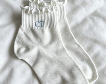 May include: A pair of white ankle socks with a ruffled trim at the top. The socks have a ribbed texture and a blue embroidered "CT" on the cuff. The socks are displayed on a white, textured surface.