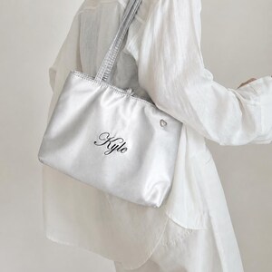 Puede incluir: Un bolso tote metálico plateado con dos correas para el hombro, que presenta el nombre Kyle bordado en letras cursivas negras en la parte frontal y un pequeño dije plateado en forma de corazón sujeto en la esquina superior derecha del exterior.