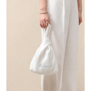 Könnte beinhalten: Eine weiße Satintasche mit geknotetem Griff. Auf der Tasche ist das Wort "Bridesmaid" in hellblauer Schrift gestickt. Die Tasche wird von einer Person gehalten, die ein silbernes Armband und einen Ring sowie eine weiße Hose trägt.