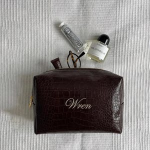 Puede incluir: Un neceser marrón oscuro texturizado con el nombre "Wren" bordado en blanco. La bolsa tiene cremallera y contiene un tubo de crema de manos, un pequeño frasco de perfume, un par de gafas y un pequeño producto labial.