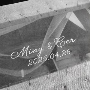 May include: Close-up of a wedding veil with the names "Ming & Cer" and the date "2025.04.26" embroidered in white script. The veil is pinned to a fabric base with small pins, creating a delicate and personalized bridal accessory.