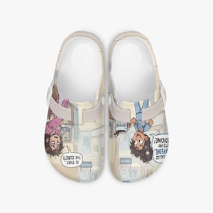 May include: A pair of beige slip-on clogs featuring a cartoon graphic of medical staff in a hospital hallway. The left shoe shows a person in pink scrubs with the text "IS THAT... THE CURE?" and the right shoe shows a person in blue scrubs holding a mug with the text "CAFFEINE" and "IT'S MY MEDICINE!"