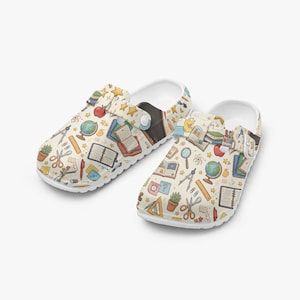 May include: White clogs featuring a cream-coloured pattern of school-themed illustrations, including books, globes, and pencils. The design also incorporates scissors and stars. The clogs have a white sole and a pivoting heel strap.