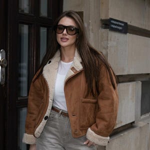 Puede incluir: Una chaqueta de ante marrón con forro y puños de borreguillo sintético color crema, sobre una camiseta blanca lisa y vaqueros grises con cinturón marrón. El conjunto se completa con unas gafas de sol de gran tamaño con cristales oscuros.