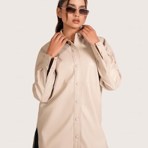 May include: A beige button-down shirt with long sleeves. The shirt has a classic collar and buttoned cuffs. The model is wearing sunglasses. The shirt is made of a smooth material and has a tailored fit.