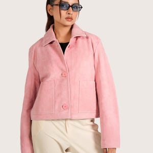 Könnte beinhalten: Hellrosa Wildlederjacke mit Kragen, zwei Vordertaschen und Knopfverschlüssen. Die Jacke wird über einem schwarzen Oberteil getragen und mit cremefarbenen Hosen kombiniert. Das Model trägt blau getönte Brillen.