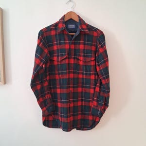 May include: A red, green, and navy plaid flannel shirt hangs on a wooden hanger. The long-sleeved shirt has two chest pockets and buttoned cuffs. The collar is lined with a blue fabric.