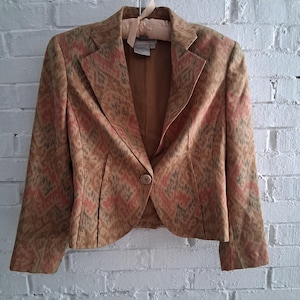 May include: A tan blazer with a patterned design in shades of pink and green. The blazer has a single button closure and is hanging on a hanger against a white brick wall. The jacket has a classic lapel and long sleeves.