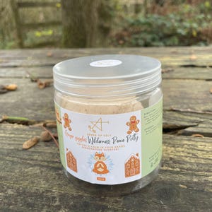 May include: A clear plastic jar with a white lid, filled with a light brown substance. The jar has a label with the text "Ginger giggles Wilderness Peace Putty" and gingerbread-themed illustrations. The jar is sitting on a wooden surface.