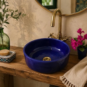 May include: A round, deep blue ceramic vessel sink with a polished gold-toned drain, paired with a matching gold-toned gooseneck faucet. The sink is positioned on a rustic wooden surface, accompanied by a small soap bar and a potted plant.