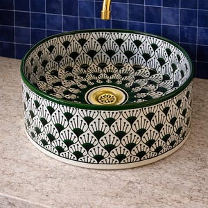 May include: A decorative ceramic vessel sink with a white background and a repeating pattern of dark green fan-like shapes. The rim is green, and the drain is gold. The sink is mounted on a light-colored countertop with a blue tile background.