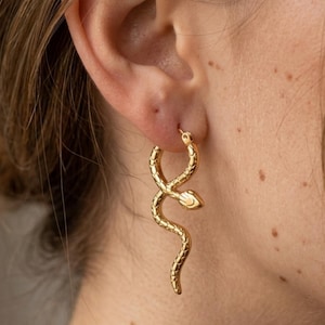 May include: Gold-colored snake hoop earrings. The earrings are worn on the ear and feature a detailed snake design. The snake's body curves and twists, with a small snake head at the bottom of the hoop. The earrings are a stylish accessory.