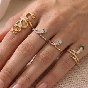 May include: Four snake-shaped rings are shown on a hand. One is gold with a textured design, the others are gold or silver with small, sparkling stones and green accents. A unique jewellery set.