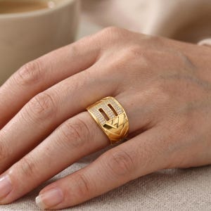 May include: A gold-colored ring with a rectangular face, set with small, clear stones. The ring has a geometric design with chevron and rectangular patterns. The ring is on a person's finger.