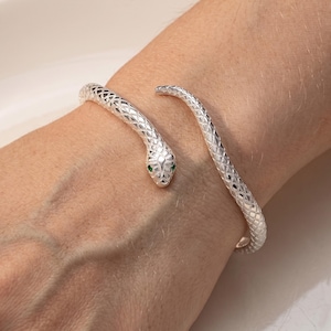 Puede incluir: Una pulsera de serpiente plateada con un patrón texturizado. La cabeza de la serpiente presenta pequeños ojos verdes. La pulsera se envuelve alrededor de la muñeca, creando un accesorio elegante y único.