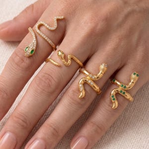 May include: A hand displays several gold snake rings. Some rings feature clear and green gemstones. The snake rings are in various designs, including a wrap-around style and a coiled design.