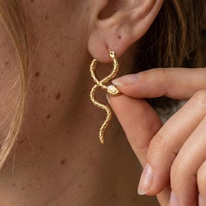 May include: Gold-colored snake hoop earrings. The earrings feature a detailed snake design with a textured surface. The snake's head is visible, and the body curves in an elegant shape. The earrings are worn on the ear.