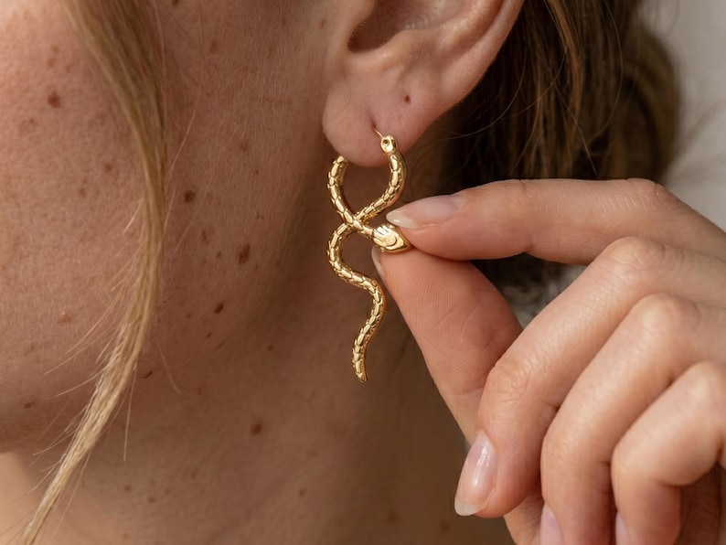 May include: Gold-colored snake hoop earrings. The earrings feature a detailed snake design with a textured surface. The snake's head is visible, and the body curves in an elegant shape. The earrings are worn on the ear.