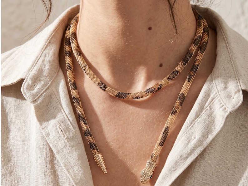 May include: A gold-toned snake necklace with a brown spotted pattern. The necklace is draped around the neck and has a pointed end with small, sparkling embellishments. The jewelry is worn over a light-colored button-down shirt.