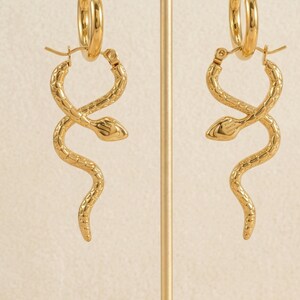 May include: A pair of gold-colored hoop earrings. Each earring features a snake design, with the snake's body wrapping around itself. The earrings are displayed on a gold-colored stand against a neutral background. The earrings are made of a shiny metal.