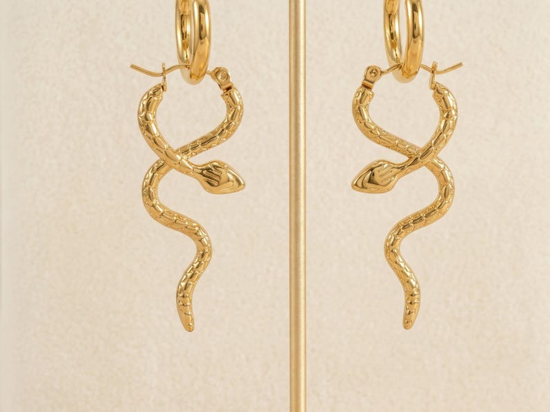 May include: A pair of gold-colored hoop earrings. Each earring features a snake design, with the snake's body wrapping around itself. The earrings are displayed on a gold-colored stand against a neutral background. The earrings are made of a shiny metal.