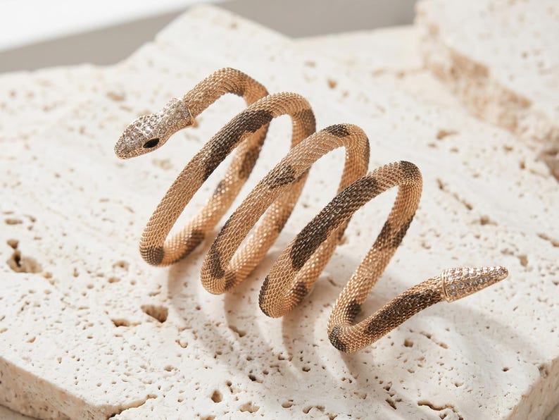 May include: A gold-toned snake-shaped bracelet with a coiled design. The snake's head and tail are detailed with small, sparkling embellishments. The body of the bracelet features a pattern of brown spots on a gold background. The jewelry is displayed on a textured, light-colored surface.