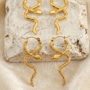 May include: Gold-colored snake hoop earrings. The earrings are in the shape of a snake, with a textured surface. The earrings are displayed on a light-colored stone surface, with a soft, cream-colored fabric in the background.