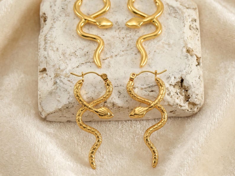 May include: Gold-colored snake hoop earrings. The earrings are in the shape of a snake, with a textured surface. The earrings are displayed on a light-colored stone surface, with a soft, cream-colored fabric in the background.
