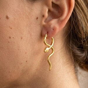 May include: A gold-colored earring in the shape of a snake is worn on a person's ear. The snake's body curves and twists, with a detailed head. The earring is a hoop style, and the snake's body hangs down. The earring is a piece of jewelry.