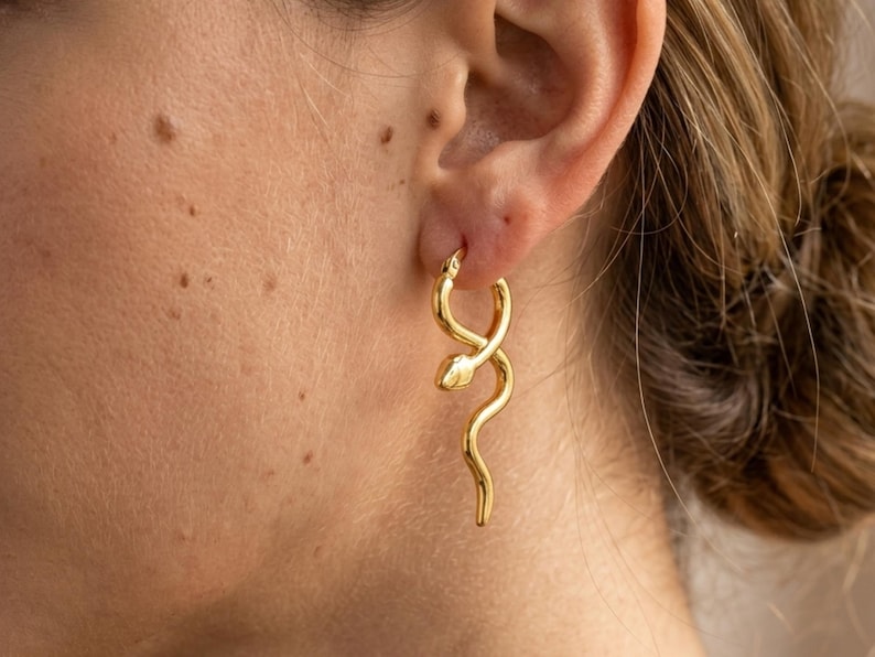 May include: A gold-colored earring in the shape of a snake is worn on a person's ear. The snake's body curves and twists, with a detailed head. The earring is a hoop style, and the snake's body hangs down. The earring is a piece of jewelry.
