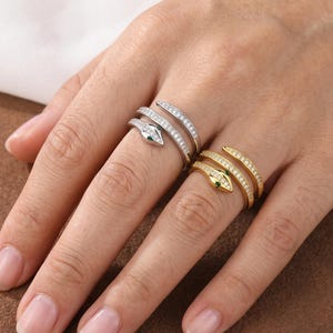 Puede incluir: Dos anillos con forma de serpiente, uno plateado y otro dorado, se muestran en una mano. El anillo plateado presenta una cabeza de serpiente con un ojo de piedra verde y está adornado con pequeñas piedras transparentes. El anillo dorado tiene un diseño similar.