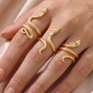 May include: Gold snake rings. The rings have detailed snake designs, with the snake's head and body wrapping around the finger. The rings are shown on a hand, showcasing the jewellery's design and texture. The rings are a statement piece.
