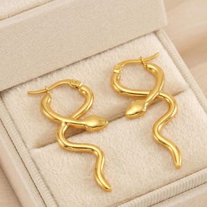 May include: A pair of gold-colored snake-shaped earrings. The earrings are in a box with a cream-colored interior. The earrings have a hoop design and a snake head at the top. The snake's body curves downward.